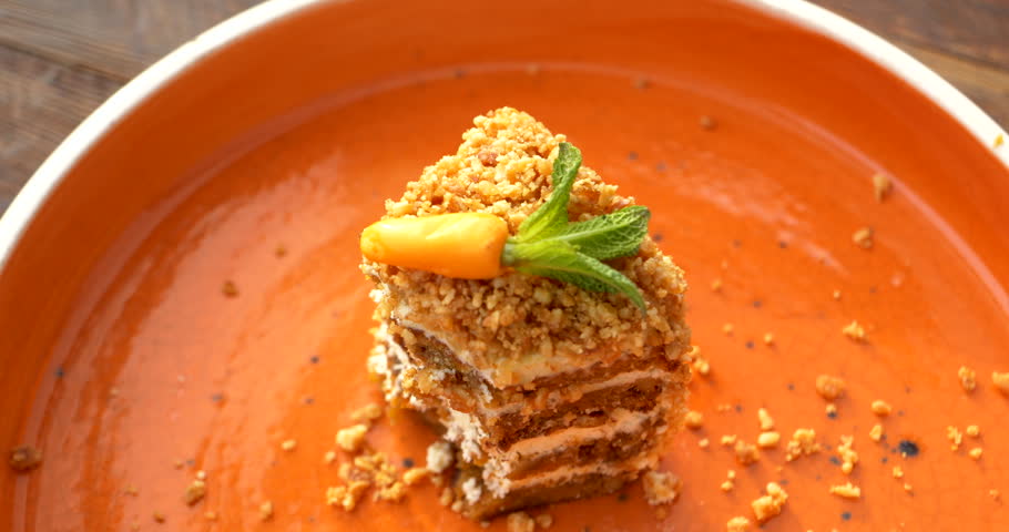 Overhead closeup shows a hand carefully using a fork to cut a soft bite from a small layered carrot cake coated in crunchy nut crumbs remove mint garnish and on a spacious glossy orange plate