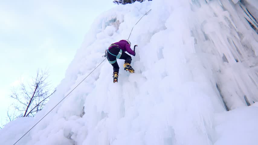 Low angle view of an alpinist woman ice climbing a steep frozen mountain wall, showcasing extreme outdoor adventure, winter sports, mountaineering, and determination.