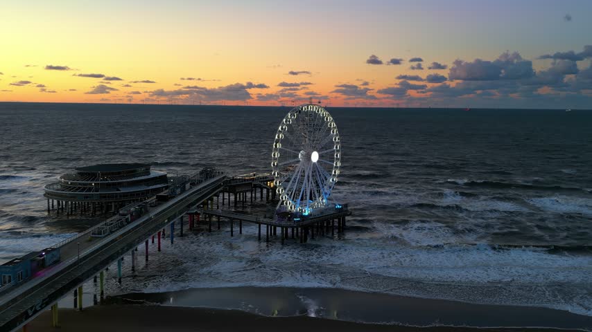 Aerial view of brightly lit ferris wheel standing on Scheveningen pier in Netherlands at sunset. Cinematic drone footage captures waves crashing against wooden structures.