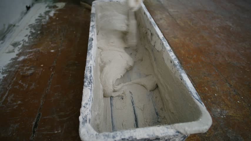 Professional worker hand mixing finishing plaster compound putty knife in plastic trough, preparing material for drywall repairs, home renovation on wooden floor. Worker mixing plaster putty in trough