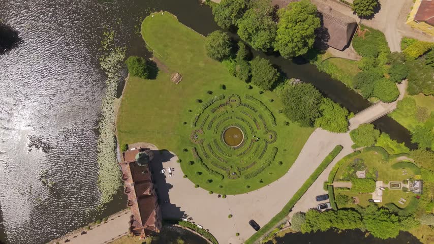 A high aerial perspective reveals the full Egeskov Castle estate, showcasing its geometric gardens, lakes, paths, and surrounding green countryside.