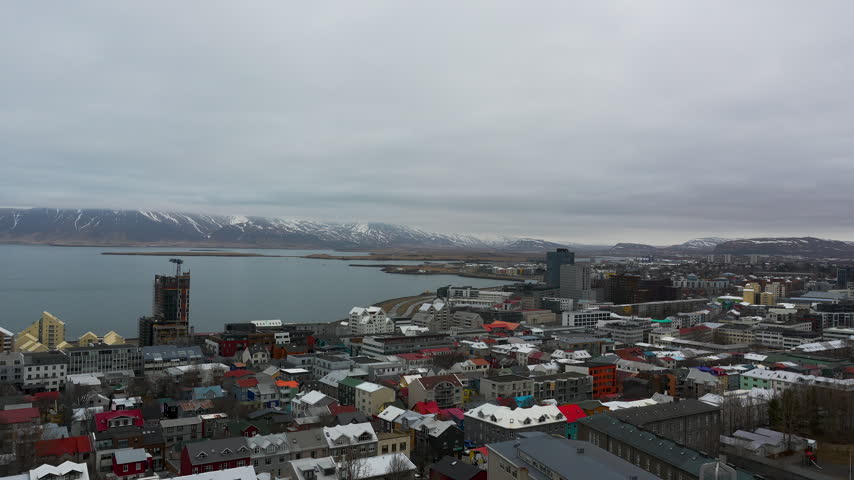A highangle aerial perspective of Reykjavik, Iceland, showcasing colorful rooftops, the bay, and distant snowy mountains under a moody overcast sky. Perfect for travel and urban planning content.