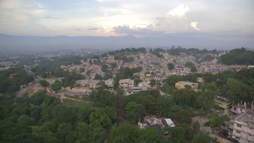 Panoramic view overlooking lush green trees and densely packed residential buildings in Port-au-Prince, Haiti. Tropical urban sprawl with moody mountains and hazy sunset skies.