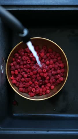 A bowl of fresh raspberries is gently rinsed under running water in a sleek black sink