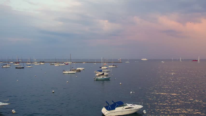 Aerial drone view of numerous sailboats and yachts anchored on Lake Michigan near a Chicago breakwater lighthouse under a soft pastel sunset sky.
