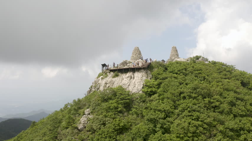 Tourists gather on a mountaintop viewing deck beside tall stone cairns above green forested slopes as a drone flies past.