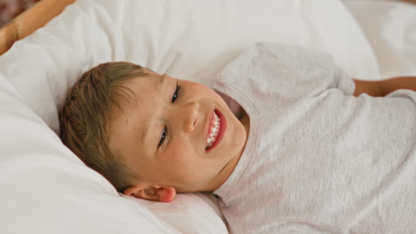 Boy lying on bed reaching toward scattered toy blocks on wooden floor, older sibling leaning over toddler in a domestic room inside a building; calm curiosity.