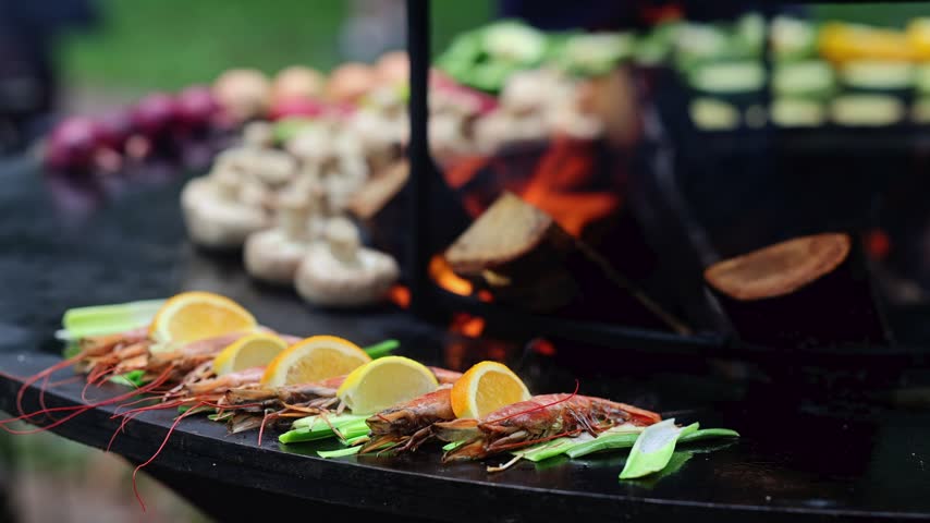 Tiger prawns grilling on round barbecue grill with fire, lemon and rosemary. Seafood barbecue cooking outdoor. Grilled shrimp with vegetables on hot charcoal flame grill closeup.