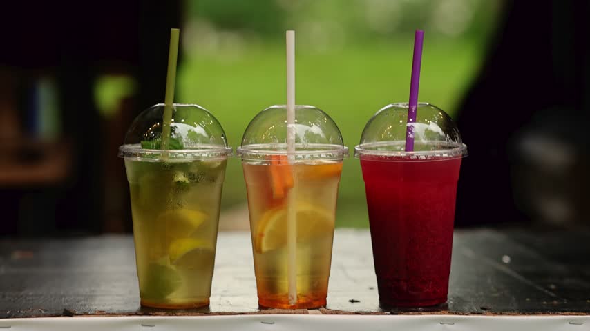 Three plastic cups with cold summer drinks with straws, lemonade with lime and mint, citrus drink and berry smoothie on table in green park. Refreshing beverages for picnic and street food concept.