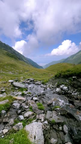 Mountain stream flows over gray rocks through a green valley in the Fagaras Mountains near Transfagarasan in Romania beneath white clouds and blue sky