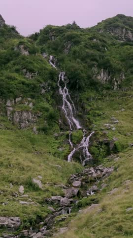 Mountain stream flows over gray rocks through a green valley in the Fagaras Mountains near Transfagarasan in Romania beneath white clouds and blue sky