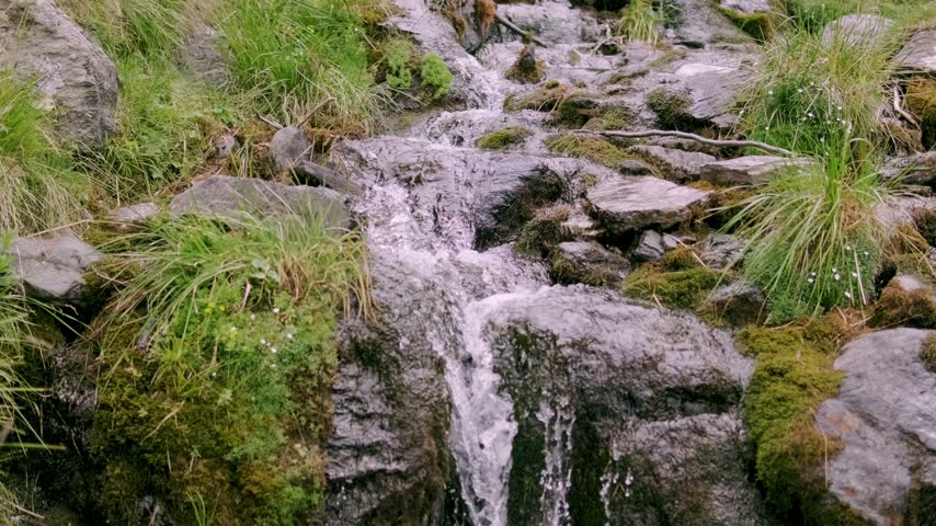 Mountain stream flows over gray rocks through a green valley in the Fagaras Mountains near Transfagarasan in Romania beneath white clouds and blue sky
