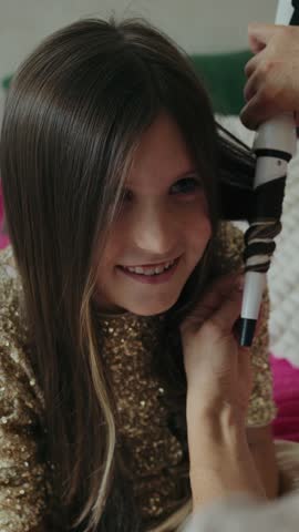 Vertical video of a cheerful little girl in a sparkly gold dress getting her hair curled with a curling iron for a festive celebration at home.