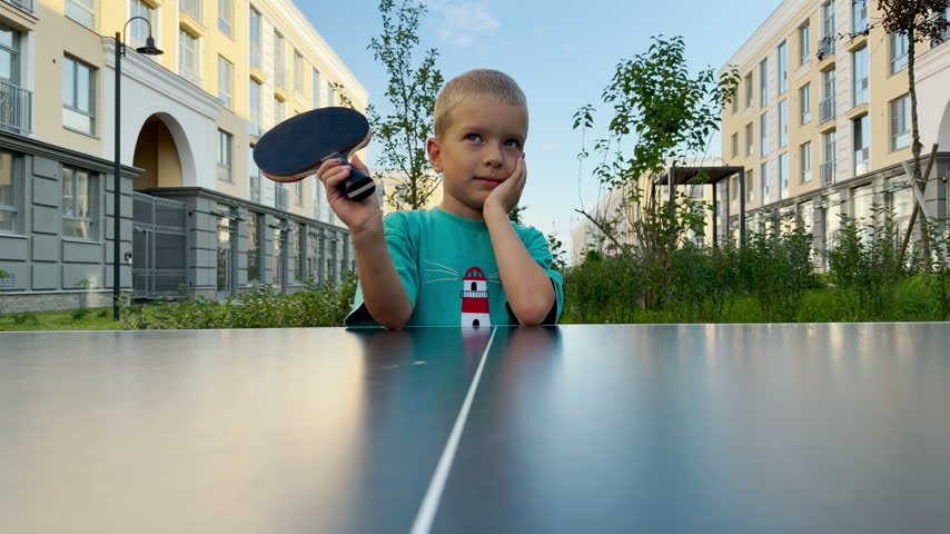 A joyful young boy rests his head and smiles while holding a ping pong paddle. He leans on an outdoor table tennis table with delight. Modern buildings frame the happy courtyard.