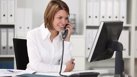 Young woman laughing on the phone sitting at her desk in an office behind her computer