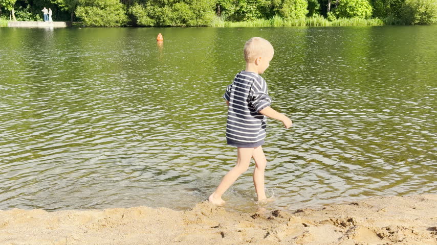 A joyful young boy wades and splashes through the water with pure happiness. He plays along the sandy shore at Kolonistsky Pond Beach in Pushkin, Saint Petersburg.