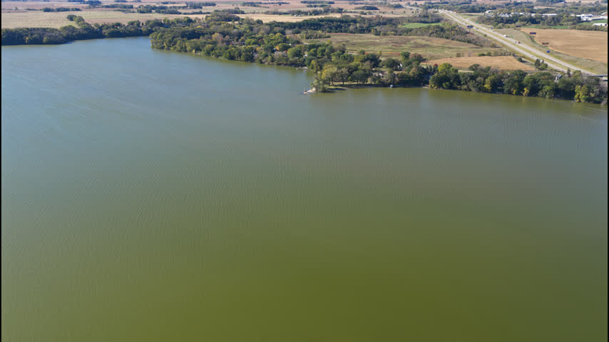 Large lake bordered by autumn woodland and farmland near Albert Lea as cars drive along highway, Minnesota, aerial establishing of Myre-Big Island State Park