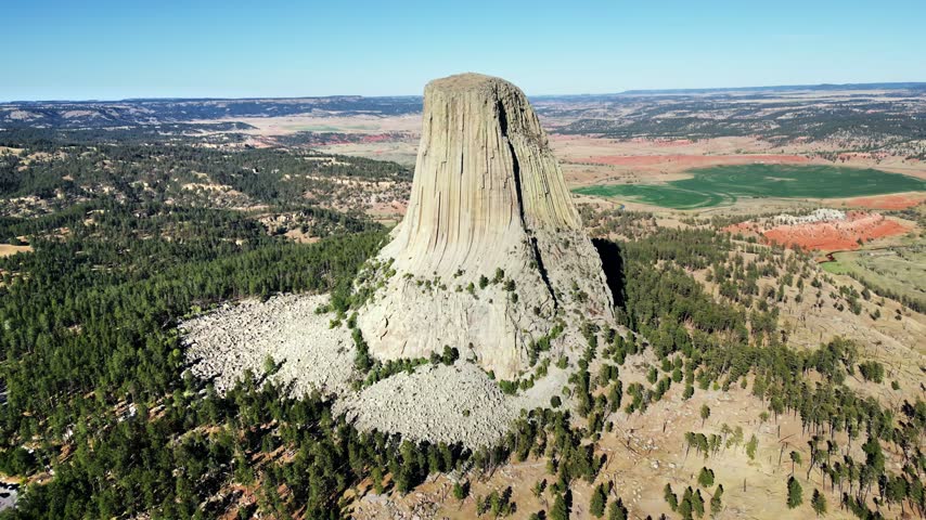 Dynamic aerial motion over devils tower national monument, revealing its striking geological formation, lush green forests, and distant red rock landscapes under a clear blue sky, perfect for cinematic travel and nature documentaries.