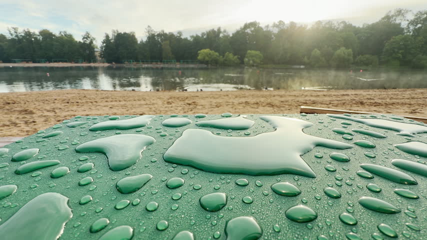 Raindrops rest on a green surface at a lake beach in Pushkin, Saint Petersburg. Soft light shines across the quiet water. The tranquil scene brings peaceful comfort.