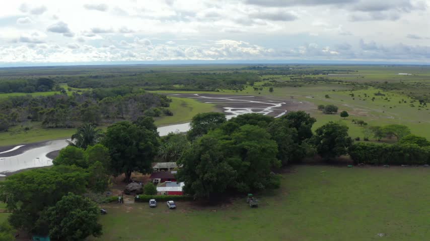 Rural ranch house shaded by large trees with parked cars, beside a winding river across the Llanos savanna in Orinoquia, Colombia, dolly left aerial shot