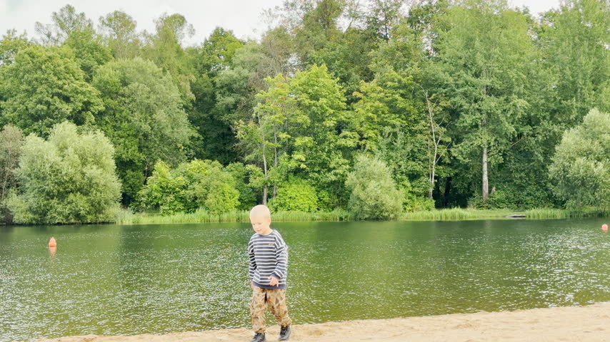 A young boy squats playfully on the sandy shore by the pond in Pushkin, Saint Petersburg. This calm scene feels peaceful, cozy, and full of innocent wonder.