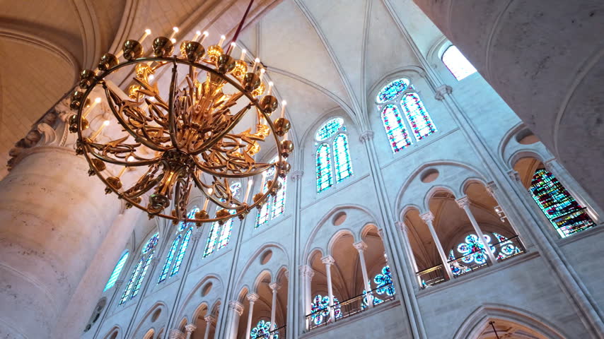 Golden cathedral chandelier hangs before tall stained glass windows inside Notre-Dame de Paris in a slow upward tilt.