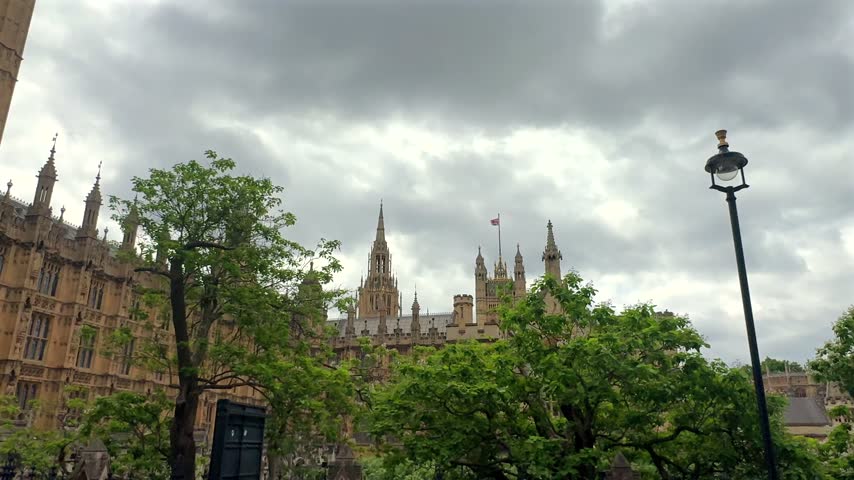 Panoramic view of the Palace of Westminster rising behind lush spring trees beneath dramatic storm clouds, showcasing ornate Gothic government architecture
