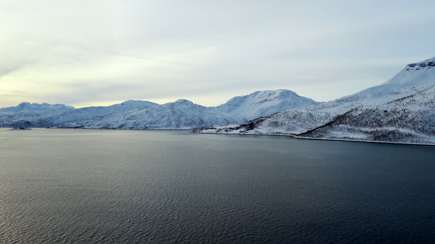 Snow-covered Arctic mountains rising beyond calm fjord waters, serene peaceful landscape backdrop of Tromso, Norway, drone