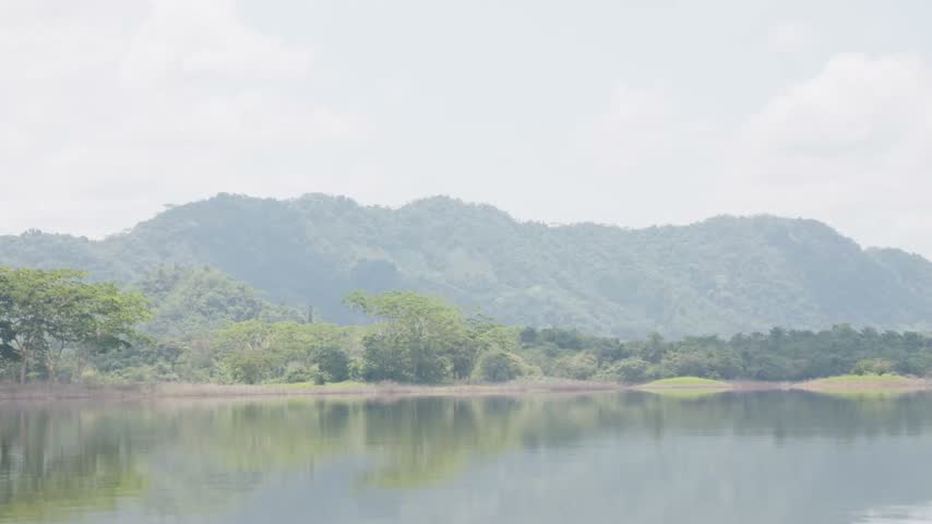 Static medium wide shot frames a hazy mountain range beyond calm water and trees