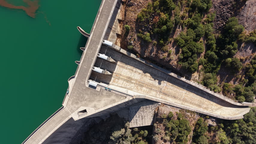 Top-down view of a large concrete dam with white turbine housings beside a turquoise reservoir, flanked by rocky cliffs and green forest