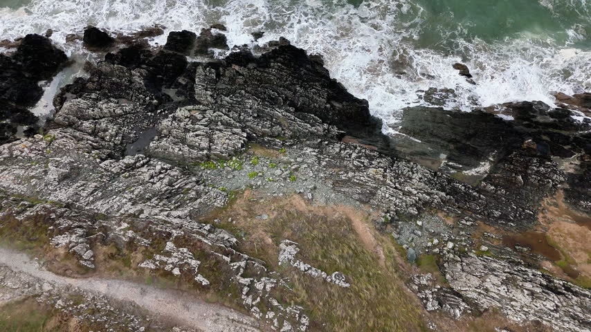 Aerial footage of historic Llanddwyn Island on the coast of Anglesey, Wales, showing large waves crashing against the rocky shoreline and historic buildings surrounded by rugged coastal scenery.