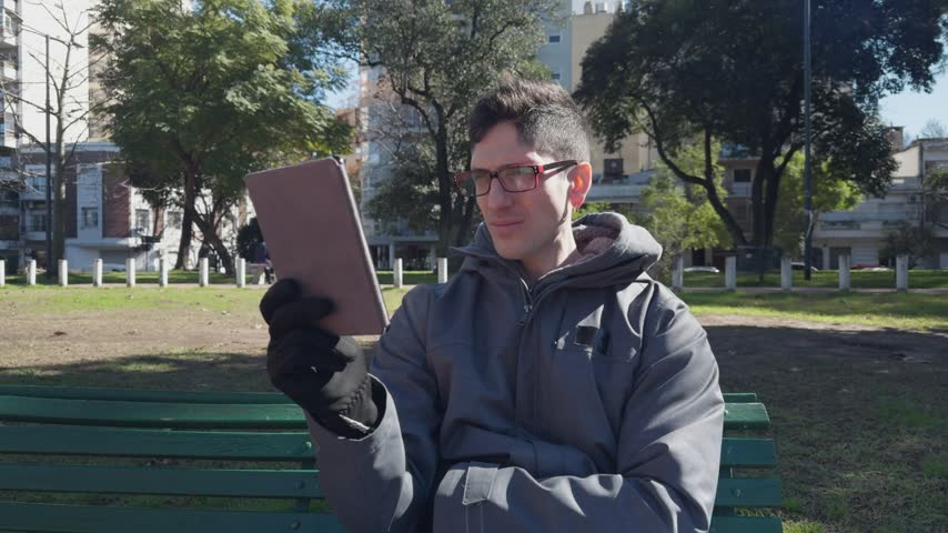 Millennial man with glasses and earphones having a video conference on his tablet in a city park