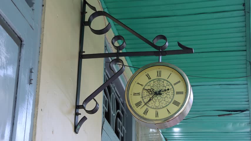 Vintage clock hangs beside a door. Black wrought iron curves above it. Clock face has Roman numerals and ornate details. Teal corrugated roof provides a backdrop. Light casts a soft glow on the scene