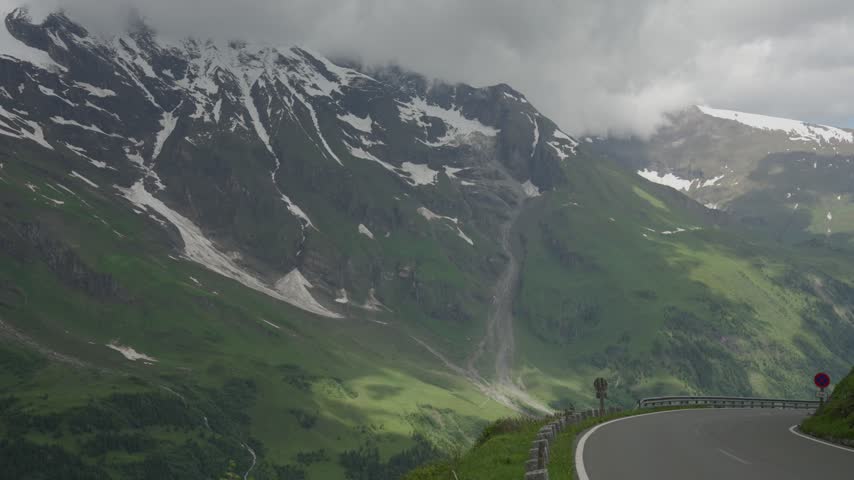 Low clouds hang over snow-streaked Austria Alps as a curving mountain road hugs the steep green slope.