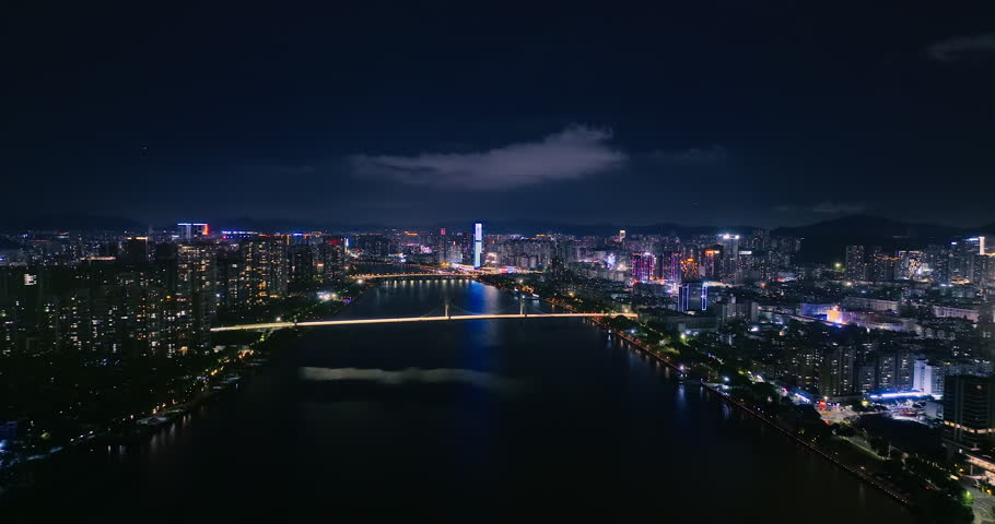 Aerial view of an illuminated city skyline at night, featuring a wide river crossed by bridges, glowing skyscrapers, and dark surrounding mountains.