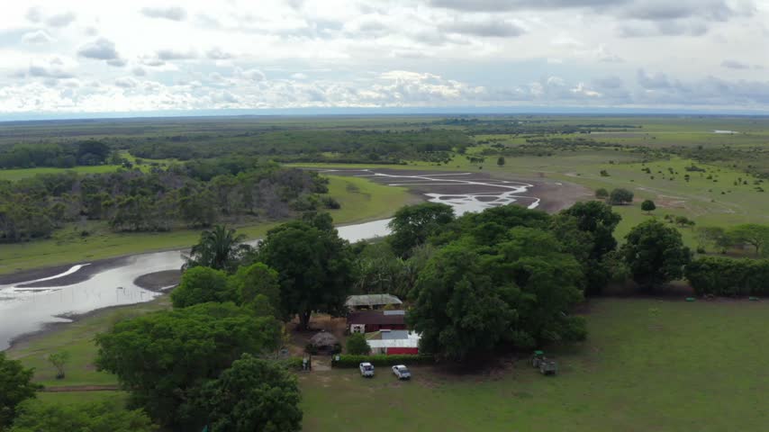 Rural ranch house shaded by large trees with parked trucks, beside braided wetland channels on the green savanna in Orinoquia, Colombia, orbit aerial shot.