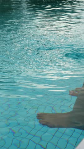 Close-up of a woman gently moving her bare feet in a swimming pool while sitting on the tiled edge. Ripples spread across the clear blue water, creating a relaxed summer leisure scene.