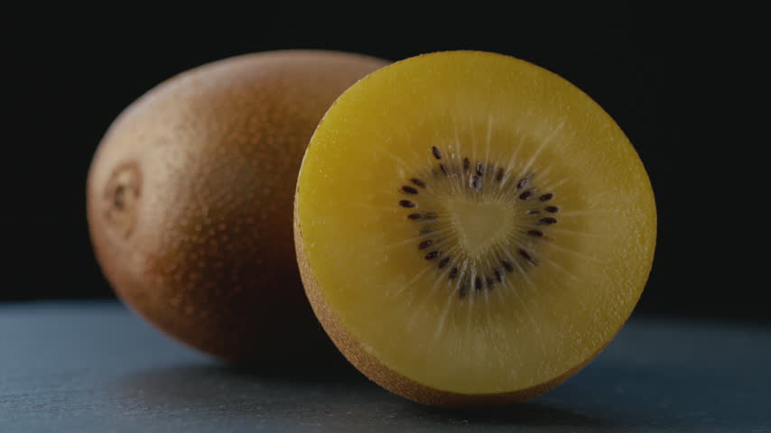 Macro shot of a ripe golden kiwi cut in half with a heart-shaped core, rich juicy pulp and seeds on a dark background with a blurred whole kiwi for healthy eating, gourmet food, and culinary content.