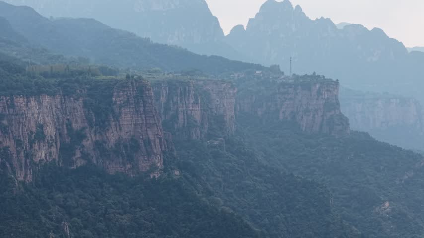 Cinematic aerial footage of majestic mountains in Henan, China. An isolated settlement on towering cliffs, deep green valleys, and hazy peaks offer an epic natural backdrop for films.