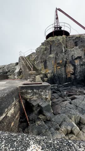 Abandoned industrial concrete pier with an old rusty crane on a rugged rocky coastline of a dark moody ocean. and man fishing for ocean fish