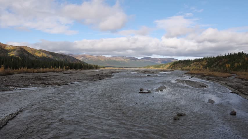 View of the river flowing through Denali National Park and Preserve with mountains and autumn foliage under a cloudy sky in Alaska, United States.