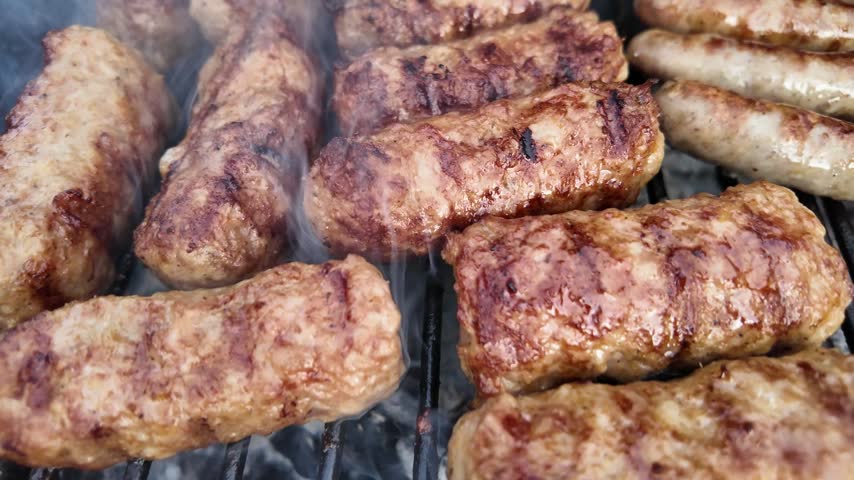 Close-up slow motion footage of traditional Romanian mici grilling over a hot barbecue, with browned meat, charred grill marks and smoke rising from the grill.