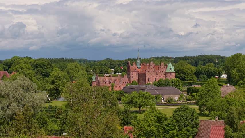 Drone footage captures Egeskov Castle on a sunny day, surrounded by lush green gardens, trees, lakes, and the peaceful Danish countryside landscape.