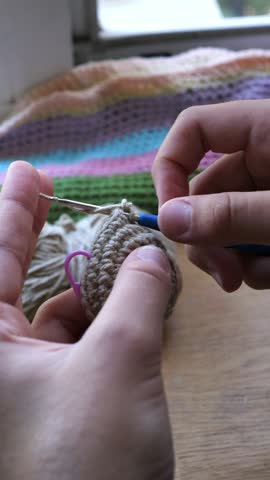 Hands crocheting a beige cotton amigurumi ball with a blue hook at a sunlit wooden table beside a striped granny blanket, cozy slow-living craft hobby