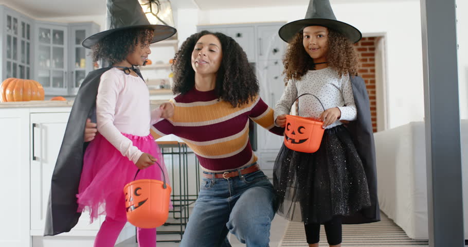 Costumed African American mother with girls fixing witch hats in kitchen then guiding with pumpkins. Family, affection, children, home, autumn, halloween, cozy