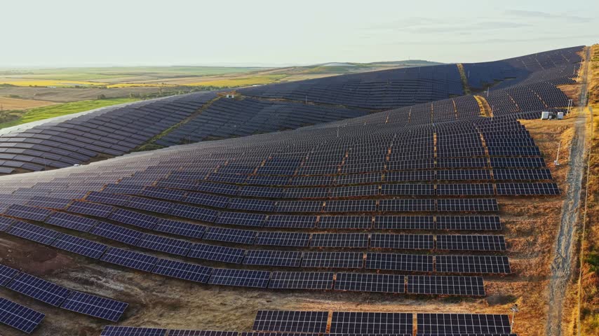 Rows of solar panels stretch across hills in a rural area. The late afternoon sun shines down, providing light for the solar farm. The landscape is open with fields visible in the distance.