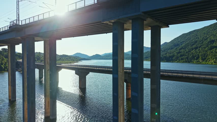 A bridge connects two banks over a river in a mountainous region. Sunlight shines down, creating reflections on the water. It is a clear day with few clouds.