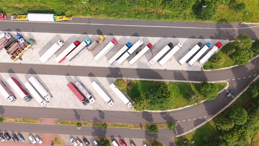 Aerial view of a busy truck parking lot showing various trucks and trailers parked in organized rows with green grass and roadways visible in the background