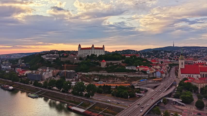 The scene shows Bratislava with the castle on a hill and the river. Cars are on the road below and buildings are visible in the city. The sky has colorful clouds as the sun sets.