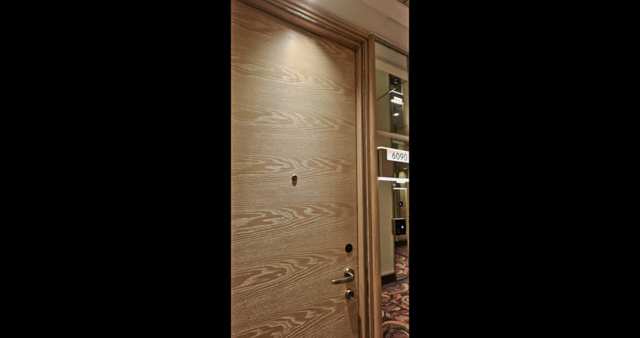 Hotel door opening from interior to a brightly lit hallway in a luxury hotel setting. Visible wood grain finish door with handle and peephole in a modern architectural building interior.